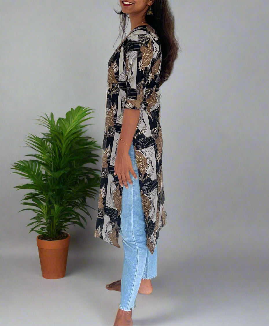Woman wearing a patterned dress and jeans standing indoors next to a green plant.